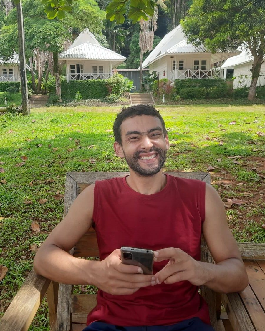 Naoufal smiling, sitting on a wooden bench in a sunny tropical garden.